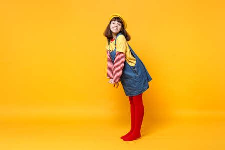 Full length portrait of beautiful girl teenager in french beret and denim sundress standing isolated on yellow wall background in studio. People sincere emotions lifestyle concept. Mock up copy spaceの写真素材