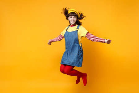 Portrait of smiling cute girl teenager in french beret and denim sundress jumping with flowing hair isolated on yellow wall background in studio. People emotions lifestyle concept. Mock up copy spaceの写真素材