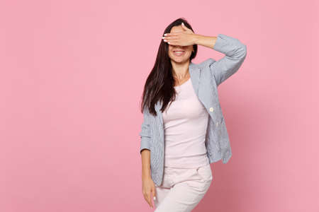 Portrait of smiling young woman in striped jacket standing, covering eyes with hand isolated on pink...