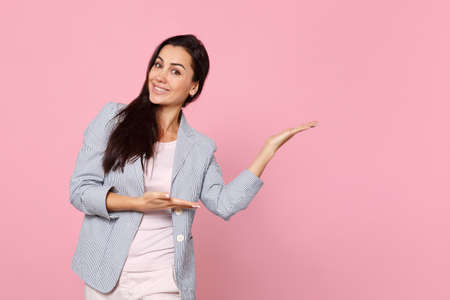 Portrait of attractive smiling young woman in striped jacket pointing hands aside isolated on pink pastel wall background in studio. People sincere emotions, lifestyle concept. Mock up copy spaceの写真素材