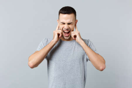 Screaming frustrated young man in casual clothes keeping eyes closed, covering ears with fingers...