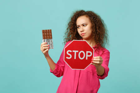 Pensive african girl in pink casual clothes holding chocolate bar, text board STOP isolated on blue turquoise wall background in studio. People sincere emotions, lifestyle concept. Mock up copy spaceの写真素材