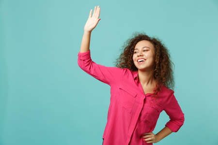 Cheerful african girl in casual clothes waving and greeting with hand as notices someone isolated on blue turquoise background in studio. People sincere emotions lifestyle concept. Mock up copy spaceの写真素材