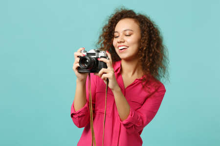 Joyful african girl in casual clothes taking picture on retro vintage photo camera isolated on blue turquoise wall background in studio. People sincere emotions lifestyle concept. Mock up copy spaceの写真素材