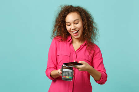Funny african girl hold mobile phone wireless modern bank payment terminal to process, acquire credit card payments isolated on blue turquoise background. People lifestyle concept. Mock up copy spaceの写真素材