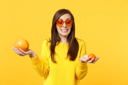 Puzzled young woman in heart glasses holding in hands fresh ripe mandarin, orange fruit isolated onÂ yellow background in studio. PeopleÂ vivid lifestyle, relax vacation concept. Mock up copy spaceの写真素材