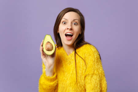 Shocked young woman in fur sweater keeping mouth open holding half of fresh ripe green avocado isolated on violet pastel background. People vivid lifestyle, relax vacation concept. Mock up copy spaceの写真素材