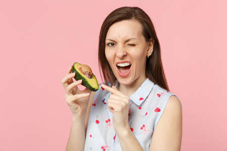 Blinking young woman in summer clothes pointing index finger on half of fresh ripe avocado fruit...