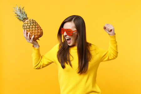 Screaming crazy young woman in funny glasses clenching fist holding fresh ripe pineapple fruit isolated onÂ yellow orange background. PeopleÂ vivid lifestyle, relax vacation concept. Mock up copy spaceの写真素材