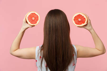 Back rear view of young woman in summer clothes hold in hands halfs of fresh ripe grapefruit isolated on pink pastel wall background. People vivid lifestyle relax vacation concept. Mock up copy spaceの写真素材