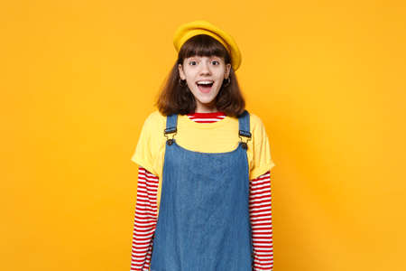 Portrait of excited cheerful funny girl teenager in french beret, denim sundress standing isolated on yellow wall background in studio. People sincere emotions, lifestyle concept. Mock up copy spaceの写真素材