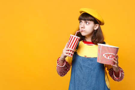 Upset girl teenager in french beret looking aside, holding plastic cup of cola or soda, bucket of popcorn isolated on yellow background. People sincere emotions, lifestyle concept. Mock up copy spaceの写真素材