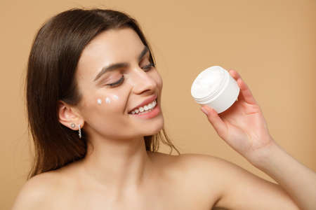 Close up half naked woman with perfect skin nude make up applying facial cream isolated on beige...