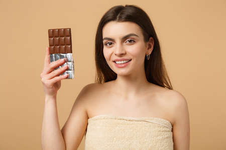 Close up half naked woman 20s with perfect skin, nude make up hold chocolate bar isolated on beige...