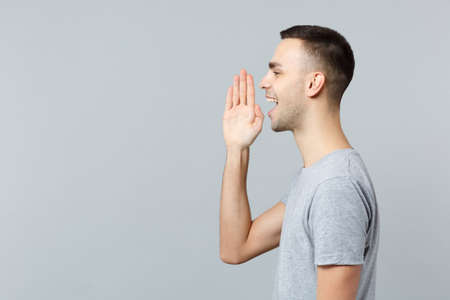 Side view of cheerful young man in casual clothes screaming with hands gesture near mouth isolated...