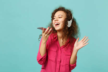 Funny african girl in casual clothes hold mobile phone, listening music with headphones isolated on...