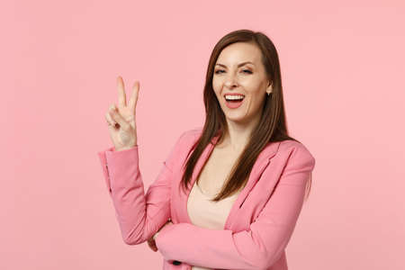 Portrait of smiling attractive young woman in jacket looking camera showing victory gesture isolated on pastel pink background in studio. People sincere emotions lifestyle concept. Mock up copy spaceの写真素材