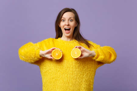 Excited young woman in fur sweater keeping mouth open holding halfs of fresh ripe orange fruit...