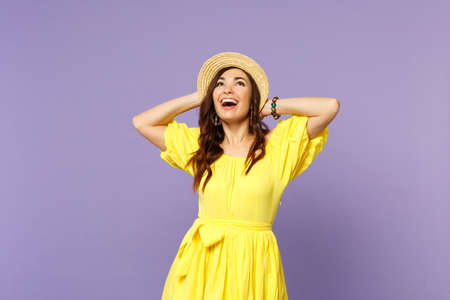 Cheerful young woman in yellow dress, hat looking up, keeping mouth open, putting hands behind head isolated on pastel violet background. People sincere emotions lifestyle concept. Mock up copy spaceの写真素材