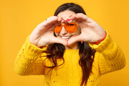 Cheerful young woman in sweater, blue trousers, sunglasses showing shape heart with hands isolated...