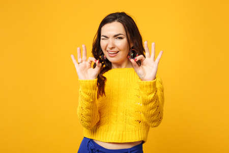 Portrait of blinking young woman in sweater, blue trousers looking camera, showing OK gesture isolated on yellow orange wall background. People sincere emotions, lifestyle concept. Mock up copy spaceの写真素材