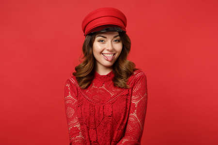 Portrait of funny cheerful young woman in lace dress, cap looking camera showing tongue isolated on bright red wall background in studio. People sincere emotions lifestyle concept. Mock up copy spaceの写真素材
