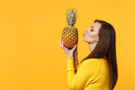Side view of young woman in casual clothes sending air kiss holding fresh ripe pineapple fruit isolated onÂ yellow orange background. PeopleÂ vivid lifestyle, relax vacation concept. Mock up copy spaceの写真素材