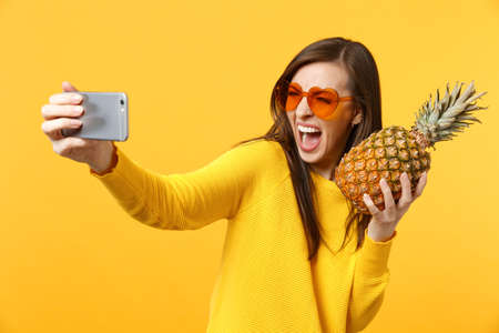 Screaming young girl in heart sunglasses hold pineapple fruit doing selfie shot on mobile phone isolated onÂ yellow orange background. PeopleÂ vivid lifestyle relax vacation concept. Mock up copy spaceの写真素材
