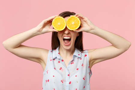 Crazy screaming young woman in summer clothes covering eyes with halfs of fresh ripe orange fruit isolated on pink pastel background. People vivid lifestyle relax vacation concept. Mock up copy spaceの写真素材