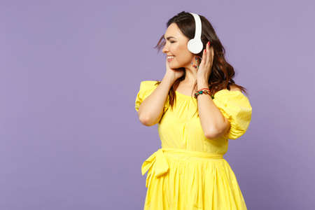 Cheerful young woman in yellow dress keeping eyes closed listen music, putting hands on headphones isolated on pastel violet background. People sincere emotions, lifestyle concept. Mock up copy spaceの写真素材