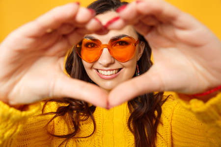 Charming young woman in sweater, blue trousers, sunglasses showing shape heart with hands isolated...