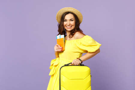 Cheerful young woman in yellow dress, summer hat hold suitcase, passport boarding pass ticket isolated on pastel violet wall background. People sincere emotions, lifestyle concept. Mock up copy spaceの写真素材