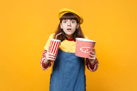 Scared girl teenager in french beret, denim sundress hold plastic cup of cola or soda, bucket of popcorn isolated on yellow background. People sincere emotions, lifestyle concept. Mock up copy spaceの写真素材