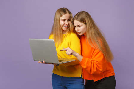 Two funny young blonde twins sisters girls in vivid colorful clothes holding, using laptop pc...