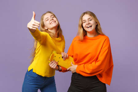 Two joyful young blonde twins sisters girls in vivid clothes showing thumb up, holding yellow...