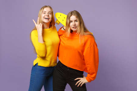 Two cheerful young blonde twins sisters girls in vivid clothes showing victory sign, holding yellow skateboard isolated on violet blue background. People family lifestyle concept. Mock up copy spaceの写真素材
