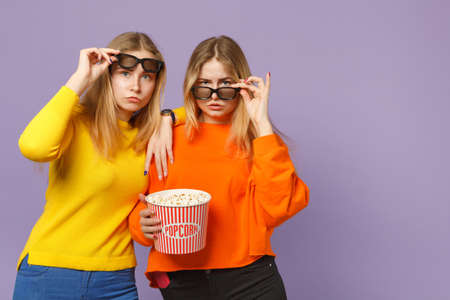 Two puzzled young blonde twins sisters girls in 3d glasses watching movie film holding popcorn isolated on pastel violet blue wall background. People family lifestyle concept. Mock up copy spaceの写真素材