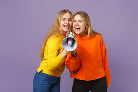 Two cheerful pretty young blonde twins sisters girls in vivid colorful clothes scream on megaphone isolated on pastel violet blue wall background. People family lifestyle concept. Mock up copy spaceの写真素材