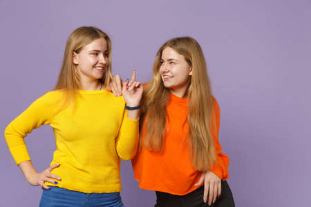 Two smiling young blonde twins sisters girls in colorful clothes looking at each other pointing index finger up isolated on violet blue background. People family lifestyle concept. Mock up copy spaceの写真素材
