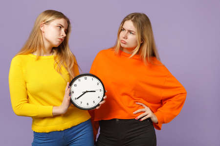 Two upset young blonde twins sisters girls in vivid colorful clothes holding round clock isolated on pastel violet blue wall background in studio. People family lifestyle concept. Mock up copy spaceの写真素材