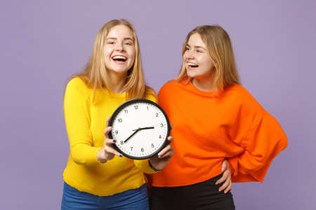 Two cheerful young blonde twins sisters girls in vivid colorful clothes hold round clock isolated on pastel violet blue wall background in studio. People family lifestyle concept. Mock up copy spaceの写真素材