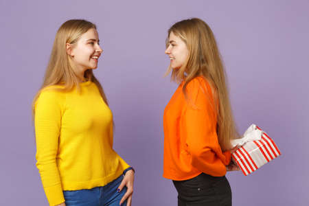 Two smiling blonde twins sisters girls in vivid clothes hold red striped present box with gift ribbon isolated on violet blue background. People family birthday, holiday concept. Mock up copy spaceの写真素材