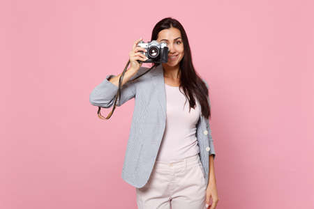 Portrait of charming young woman in striped jacket taking pictures on retro vintage photo camera isolated on pink pastel wall background. People sincere emotions lifestyle concept. Mock up copy spaceの写真素材