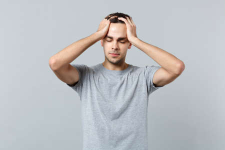 Portrait of tired young man in casual clothes keeping eyes closed, putting hands on head isolated on grey wall background in studio. People sincere emotions, lifestyle concept. Mock up copy spaceの写真素材