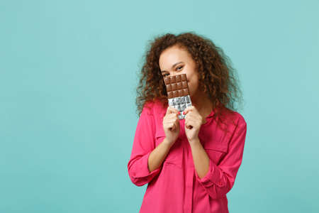 Portrait of cute african girl in casual clothes covering face with chocolate bar isolated on blue turquoise wall background in studio. People sincere emotions, lifestyle concept. Mock up copy spaceの写真素材