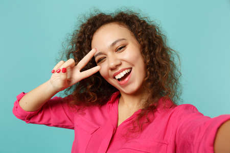 Close up selfie shot of blinking african girl in casual clothes showing victory sign isolated on blue turquoise background in studio. People sincere emotions, lifestyle concept. Mock up copy spaceの写真素材
