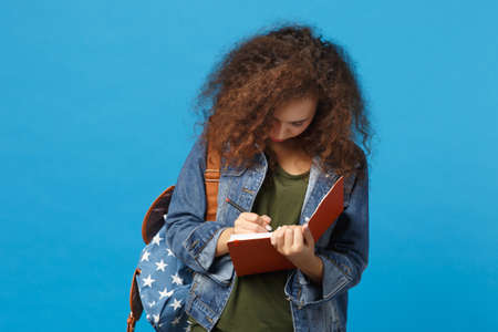 Young african american girl teen student in denim clothes, backpack hold books isolated on blue background studio portrait. Education in high school university college concept. Mock up copy spaceの写真素材
