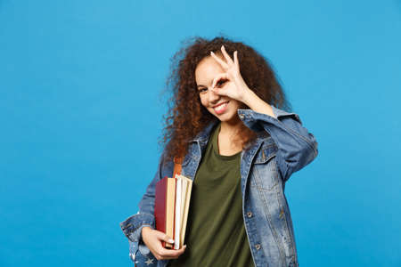 Young african american girl teen student in denim clothes, backpack hold books isolated on blue background studio portrait. Education in high school university college concept. Mock up copy spaceの写真素材