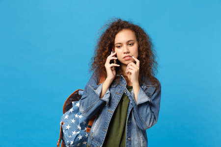 Young african american girl teen student in denim clothes, backpack hold phone isolated on blue...