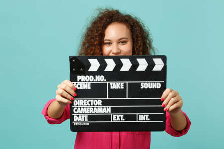 Pretty african girl in casual clothes covering face with classic black film making clapperboard isolated on blue turquoise background. People sincere emotions, lifestyle concept. Mock up copy spaceの写真素材